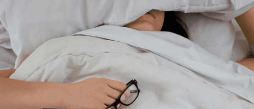 A person lying in bed under white bedding with a pillow covering their face, holding a pair of glasses in one hand, suggesting exhaustion or rest.