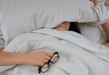 A person lying in bed under white bedding with a pillow covering their face, holding a pair of glasses in one hand, suggesting exhaustion or rest.