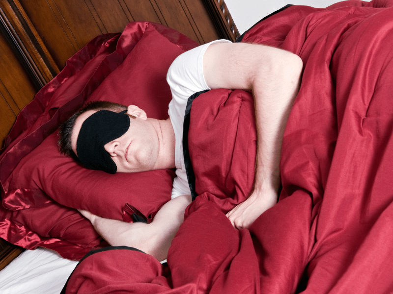 man with a black sleep mask on and sleeping in side position with head on red pillow and covered by red blanket