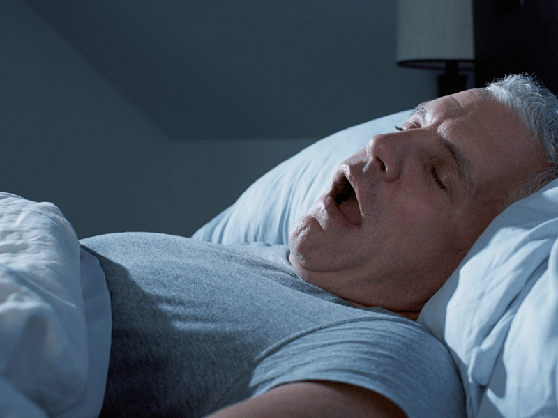 older man with white hair lying in bed and mouth open, snoring.