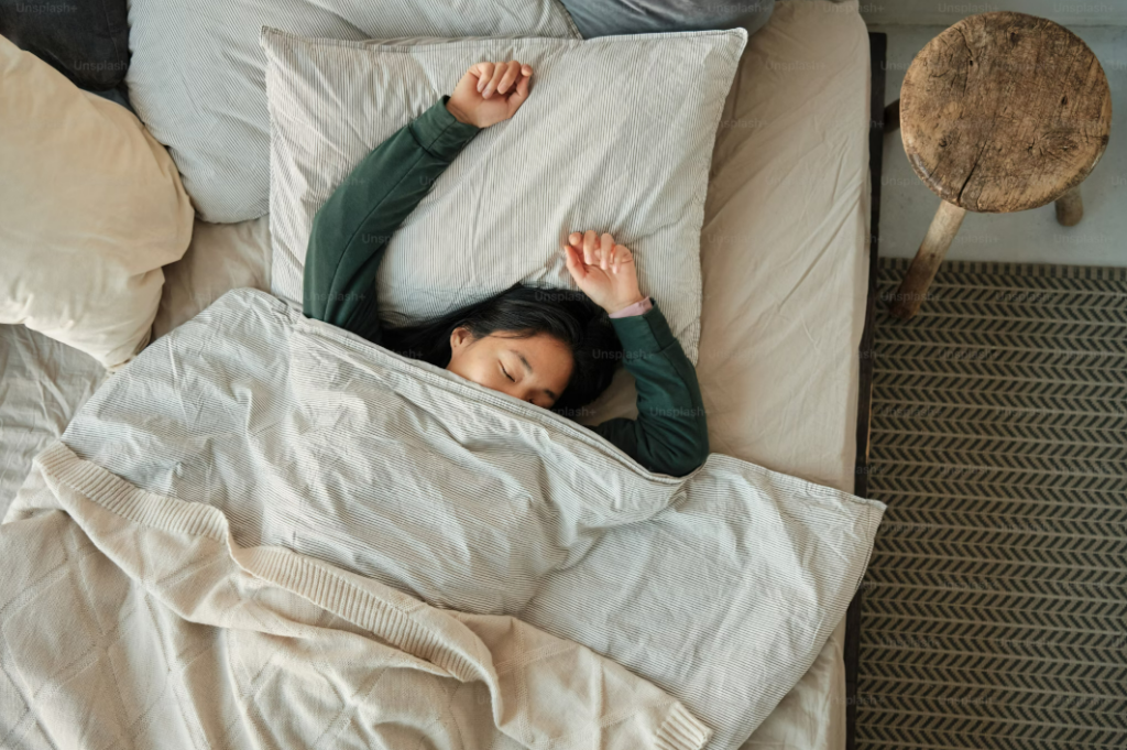 A teenager sleeping peacefully in bed, lying on a striped pillow, covered with a cozy blanket, and stretching their arms above their head in a relaxed posture.