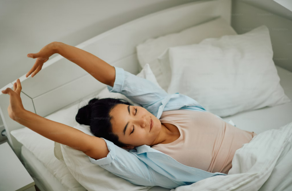 A woman waking up feeling refreshed, stretching her arms in bed with a peaceful expression, surrounded by soft white pillows and bedding, indicating a good night's sleep.