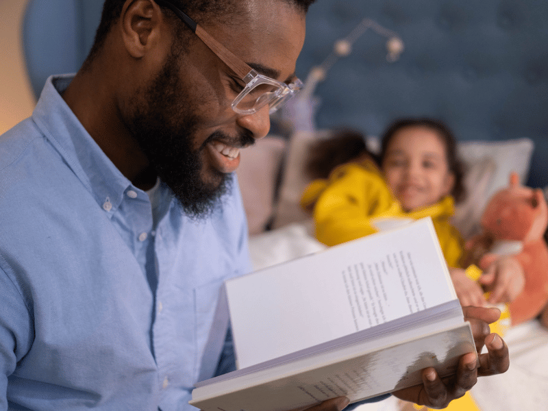 a father smiling and reading a book to his daughter in the background bedtime stories for adults