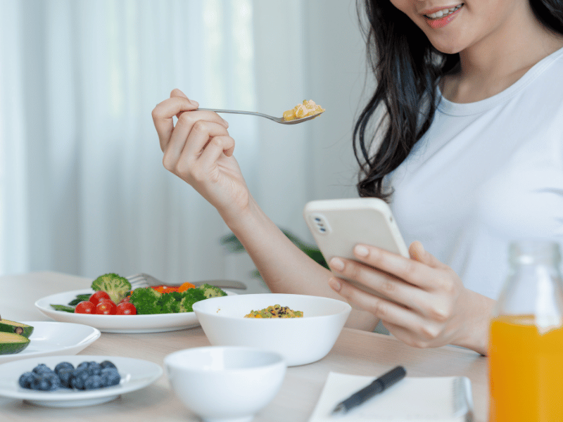 A woman at a table with a phone and a bowl of food, enjoying her meal while using her phone.