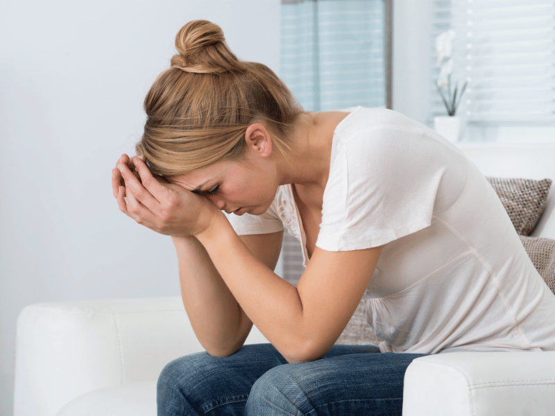 A woman sitting on a couch, resting her face in her hands, looking stressed, a factor that causes a woman to need more sleep 