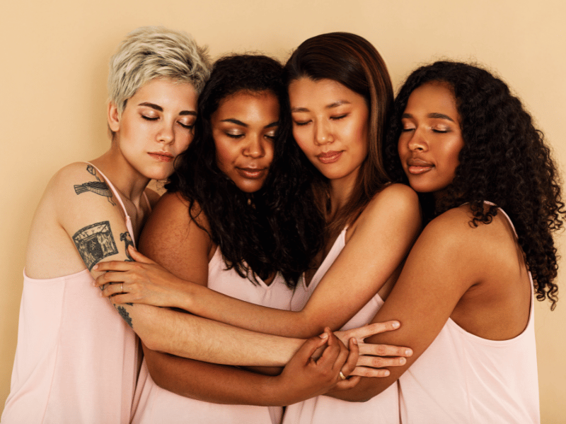  Four women in pink shirts share a warm hug, symbolizing the need for more sleep for women compared to men.