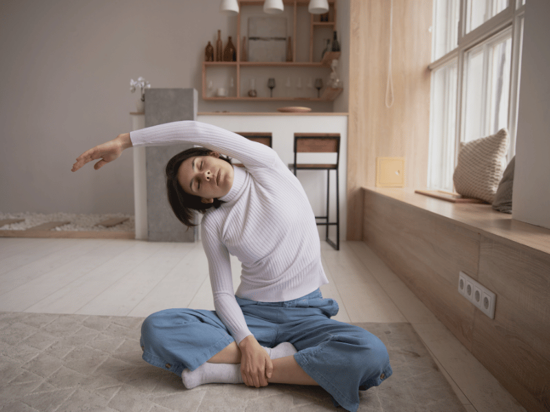 woman sitting cross-legged on the ground with eyes closed and her arm stretched out to one side as a form of quick stretch or workout